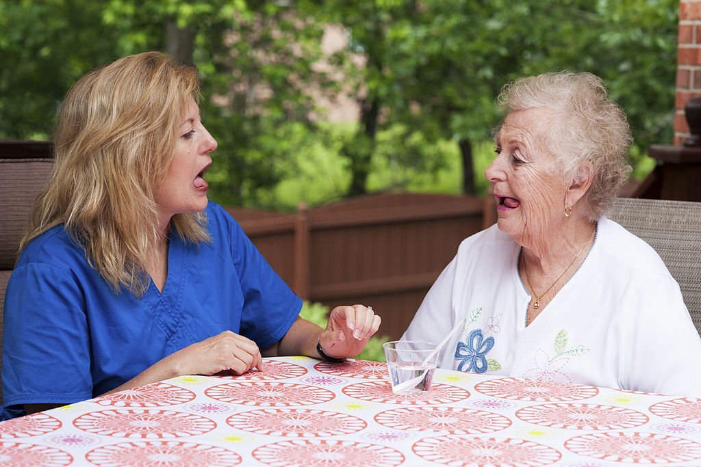 A blond speech therapist models oral motor exercises while a senior female stroke patient imitates the lateral tongue movements while sitting outdoors during a home health therapy session on a summer day